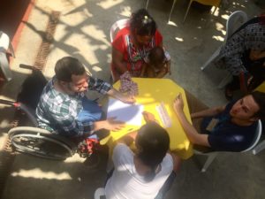 Foto composta por quatro pessoas sentada em uma mesa, são dois homens e duas mulheres o homem da esquerda esta sentado em uma cadeira de rodas a sua esquerda uma mulher segura um bebê, ambos estão discutindo.
