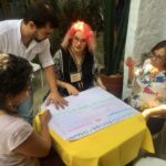 Um grupo de pessoas reunidos em volta de uma mesa, sentadas existem 3 senhoras e um homem em pé.