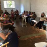 Foto de um home em pé e de costas para a foto, palestra para uma turma sentada em círculo.