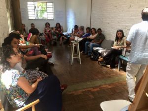 Foto de um home em pé e de costas para a foto, palestra para uma turma sentada em círculo.
