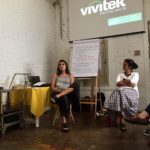 Foto composta de duas pessoas sentadas, onde uma jovem de costas para um quadro de anotações fala com um publico, a sua esquerda uma jovem a observa.