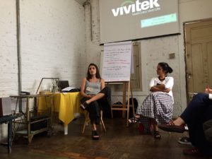 Foto composta de duas pessoas sentadas, onde uma jovem de costas para um quadro de anotações fala com um publico, a sua esquerda uma jovem a observa.