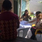 Foto de três mulheres, duas estão sentadas em uma mesa e discutindo enquanto uma mulher negra de costa para a câmera as observa.