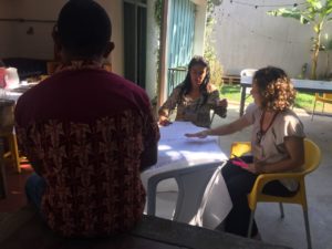 Foto de três mulheres, duas estão sentadas em uma mesa e discutindo enquanto uma mulher negra de costa para a câmera as observa.