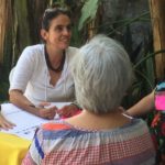 Foto composta por quatro mulheres sentadas em volta de uma mesa, uma senhora de cabelo grisalho está de costa para a foto, a sua direita uma mulher de roupa preta está sorrindo, ao lado direito dela uma mulher de branco sorrindo ao lado direito dela, uma mulher está apoiando seu braço na mesa.