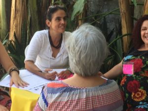 Foto composta por quatro mulheres sentadas em volta de uma mesa, uma senhora de cabelo grisalho está de costa para a foto, a sua direita uma mulher de roupa preta está sorrindo, ao lado direito dela uma mulher de branco sorrindo ao lado direito dela, uma mulher está apoiando seu braço na mesa.